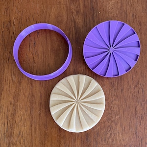May include: A purple circular cookie cutter, a matching purple circular stamp with a radial sunburst pattern, and a beige circular cookie dough piece featuring the same embossed sunburst design, all arranged on a flat surface.