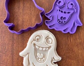 May include: A purple plastic cookie cutter and a white cookie in the shape of a cartoon ghost with a wide open mouth and big eyes.