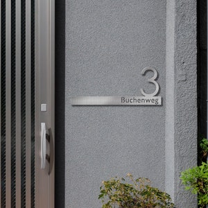 May include: A silver house number plaque with the number "3" and the text "Buchenweg" on a gray wall.
