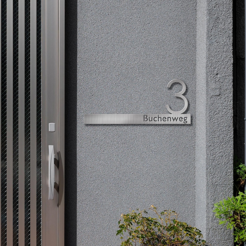 May include: A silver house number plaque with the number "3" and the text "Buchenweg" on a gray wall.