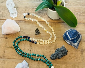 May include: A green malachite mala necklace with a white tassel, a yellow citrine mala necklace, a small gold Buddha statue, a white crystal statue, a pink rose quartz, a grey stone, and a blue lapis lazuli crystal.