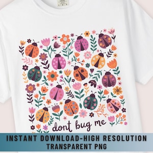 Op de afbeelding: Wit t-shirt met een kleurrijk bloemen- en lieveheersbeestjesontwerp. Het ontwerp bevat verschillende lieveheersbeestjes in oranje, roze, groen en paars, omringd door bloemen en bladeren. De tekst "don't bug me" is ook opgenomen.