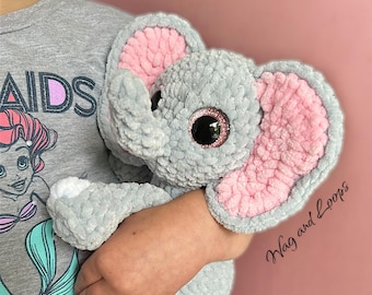 May include: A gray crocheted elephant plush toy with pink ears and large, sparkly eyes. The elephant is being held by a person wearing a gray shirt with the text "AIDS" and "HAVE FUN" on it.