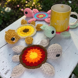 May include: A collection of handmade crochet turtle coasters in various colors, including pink, yellow, green, and brown. One coaster has a sunflower design, and another has a vibrant red, orange, and yellow pattern. A yellow mug with the text "BUZZ OFF" rests on a pink coaster.