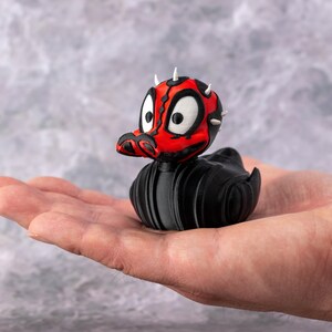 May include: A black rubber duck with a red and black face, white eyes, and small white spikes on its head. The duck is held in a person's open hand against a gray background. The duck is a novelty item.