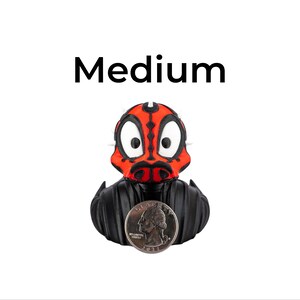 May include: A rubber ducky with a red and black face design, resembling a character from a science fiction film. The ducky has a black body and a quarter coin is in front of it. The word "Medium" is above the ducky.