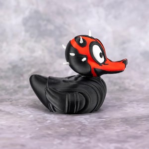 May include: A black rubber duck with a red and black face, resembling a character from a science fiction film. The duck has white horns and a detailed design. The duck is on a gray surface.
