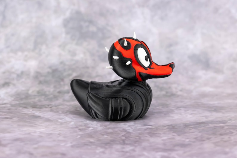 May include: A black rubber duck with a red and black face, resembling a character from a science fiction film. The duck has white horns and a detailed design. The duck is on a gray surface.