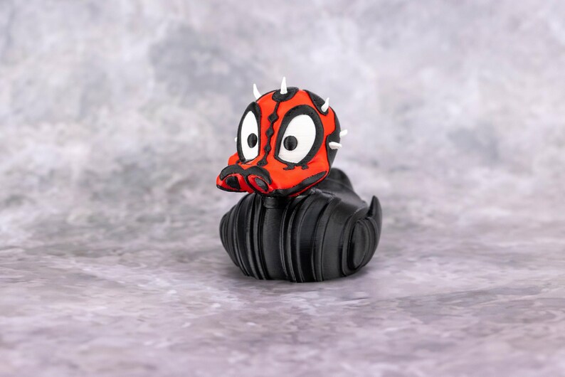 May include: A novelty rubber duck with a red and black painted head, white eyes, and small white horns. The duck's body is black. The duck is a unique collectible item.
