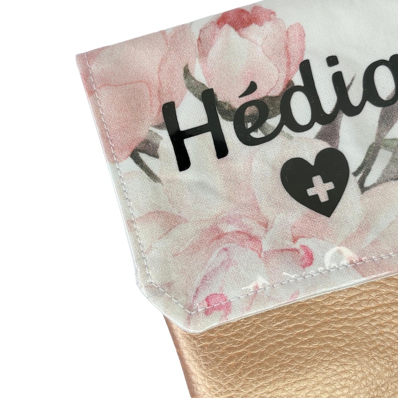May include: A white fabric pouch with a floral pattern and a gold leather bottom. The pouch has a black vinyl name tag that says "H&eacute;dia" and a black heart with a white plus sign.