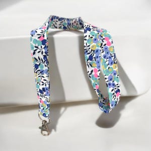 Peut inclure: Un cordon floral blanc orné de motifs floraux bleus, roses et jaunes avec des feuilles vertes. Il est doté d'un fermoir en métal argenté.