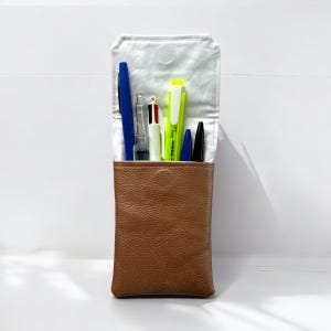 May include: A brown leather pencil case with a white fabric lining. The case is filled with pens and pencils, including a blue pen, a black pen, a red and black pen, a white pen, and a yellow highlighter.