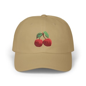 May include: A tan baseball cap featuring an embroidered design of two red cherries with green stems and leaves centered on the front panel. The cap has a structured crown, a curved brim, and a button at the top of the dome.