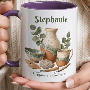 May include: A white ceramic mug with a purple interior and handle, featuring the name Stephanie, a pottery illustration with a vase, bowl, mug, and tools, and the text Happiness is handmade in dark green lettering on the front surface.