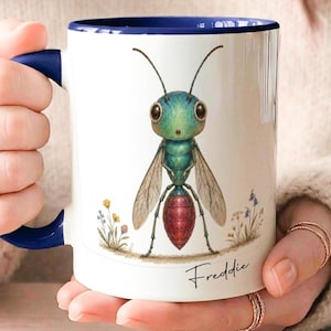 Peut inclure: Un mug en céramique blanche avec une anse et un intérieur bleu foncé. Le mug présente une illustration colorée d'un insecte avec une tête, un thorax et des ailes translucides bleu sarcelle, ainsi qu'un abdomen rouge. Des fleurs sauvages entourent l'insecte et le nom Freddie est imprimé en écriture noire sous l'illustration.