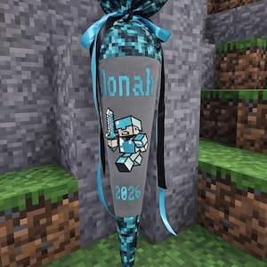 Könnte beinhalten: Eine kegelförmige Geschenktüte aus Stoff mit einem grauen Mittelteil, auf dem der Name Jonah und das Jahr 2026 in Blau gestickt sind. Die Tüte hat oben und unten ein blau-schwarzes Pixelmuster und ist mit blau-schwarzen Satinbändern verziert.