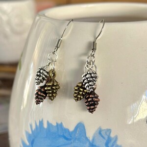 Puede incluir: Un par de pendientes de aro de plata con tres pequeños abalorios de piña. Las piñas son de color plateado, dorado y cobre.