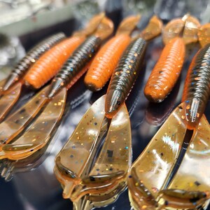 May include: A close-up of several orange and brown soft plastic fishing lures with a glittery finish. The lures have a segmented body and a forked tail.