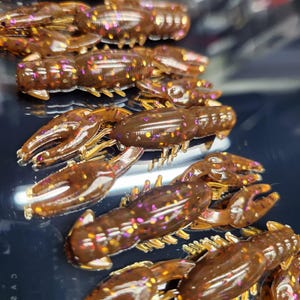 May include: A close-up of a pile of brown and gold crawfish-shaped fishing lures with glitter accents.