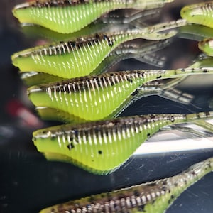 May include: A close-up of five green and black soft plastic fishing lures. The lures have a ribbed texture and a pointed tail.