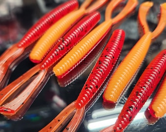 May include: A close-up of several orange and red fishing lures with black flecks. The lures have a split tail design.
