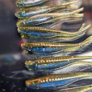 May include: A close-up of a pile of green and blue fishing lures with glitter. The lures are arranged in a stack, with the tails of the lures pointing towards the top of the image.