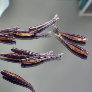 May include: A collection of purple and gold soft plastic fishing lures. The lures are shaped like small fish with a translucent body and a shimmering finish. Green glitter accents the lures.