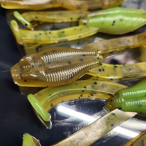May include: A close-up of several green and brown soft plastic fishing lures. The lures have a speckled pattern and are designed to resemble frogs.