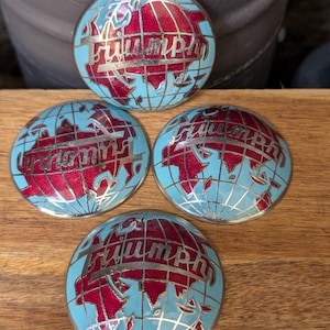 May include: Four vintage Triumph emblem badges. Each badge features a globe design with red continents, a light blue ocean, and the word "Triumph" in silver lettering. The badges are arranged on a wooden surface.