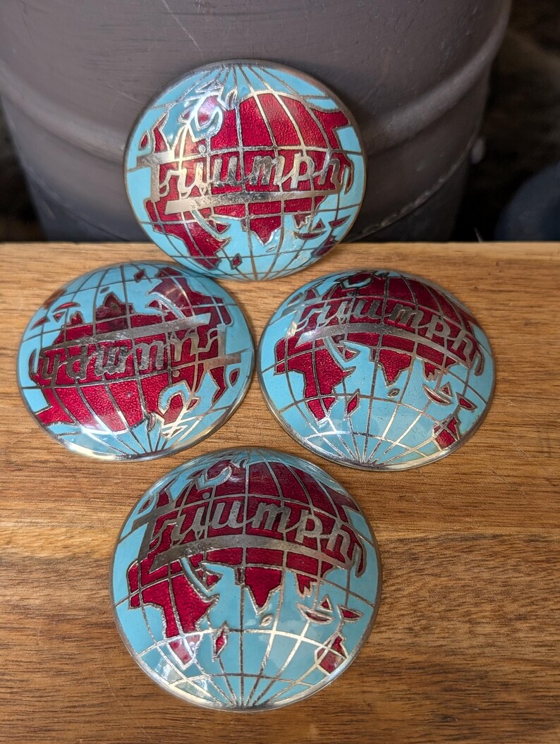 May include: Four vintage Triumph emblem badges. Each badge features a globe design with red continents, a light blue ocean, and the word "Triumph" in silver lettering. The badges are arranged on a wooden surface.