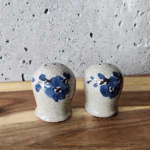 May include: A pair of ceramic salt and pepper shakers with a floral design. The shakers are white with blue flowers and green leaves.