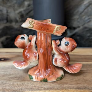 May include: A ceramic salt and pepper shaker set featuring two brown squirrels climbing a central tree trunk. A horizontal wooden-style sign is attached to the top of the trunk with the gold letters S and P clearly visible on the front surface.