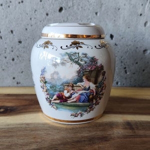 May include: White ceramic jar with a gold trim and a floral design. The jar features a painted scene of two people sitting in a garden.