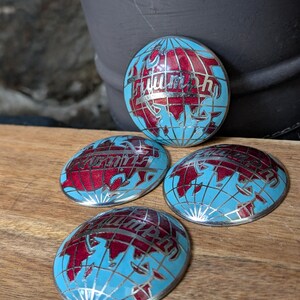 May include: Four oval-shaped Triumph emblems. Each emblem features a light blue globe design with red continents and silver accents. The word "Triumph" is written in red script across the globe. The emblems are on a wooden surface.