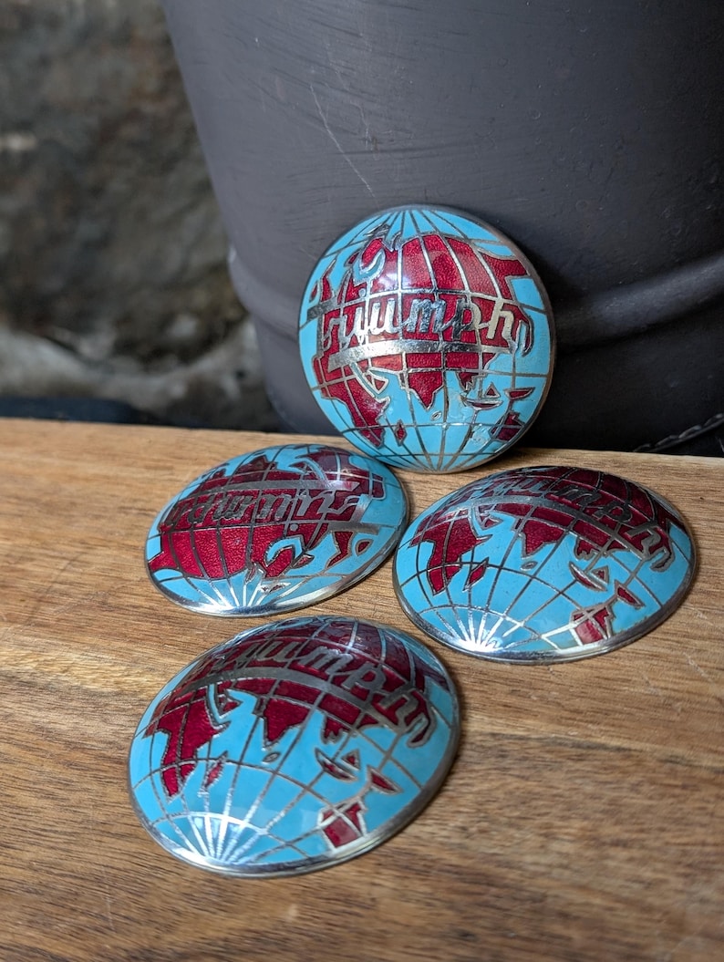 May include: Four oval-shaped Triumph emblems. Each emblem features a light blue globe design with red continents and silver accents. The word "Triumph" is written in red script across the globe. The emblems are on a wooden surface.