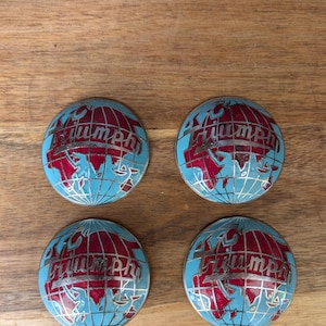May include: Four round, blue and red Triumph emblems. Each emblem features a globe design with red continents and the word "Triumph" in silver lettering. The emblems are arranged on a wooden surface.