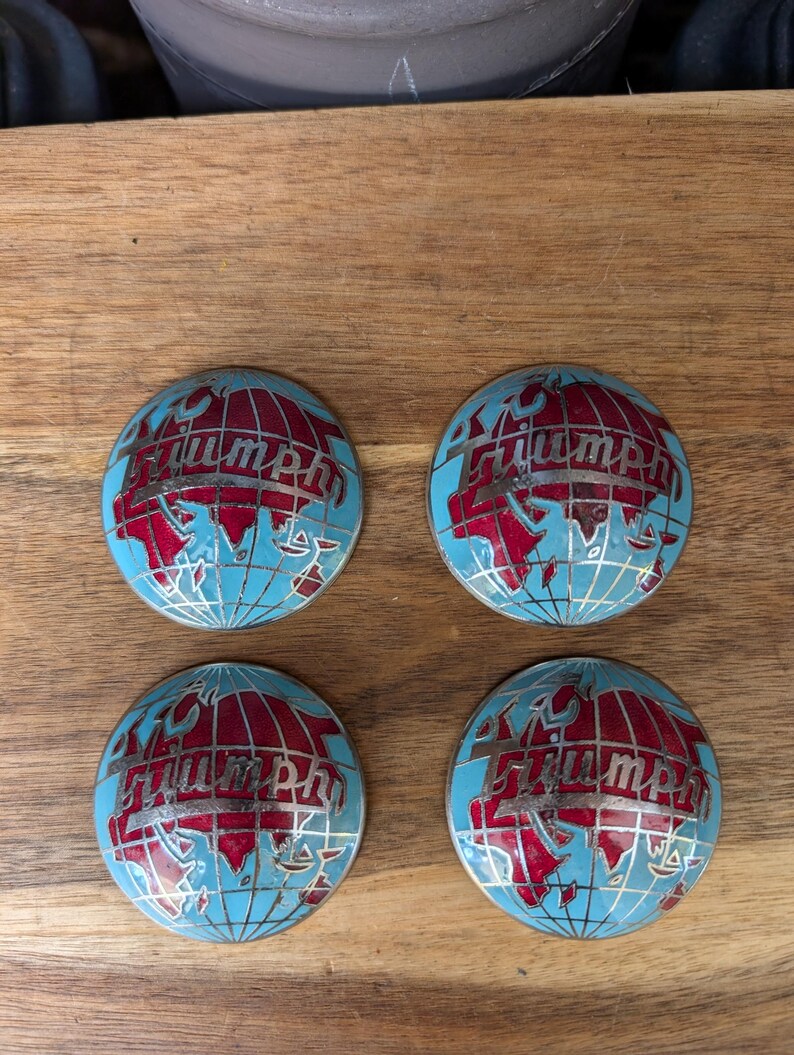 May include: Four round, blue and red Triumph emblems. Each emblem features a globe design with red continents and the word "Triumph" in silver lettering. The emblems are arranged on a wooden surface.