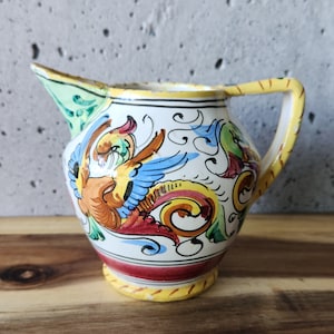 Peut inclure: Un petit pichet en céramique blanche avec un motif floral jaune, rouge et bleu. Le pichet a une anse et un bec.