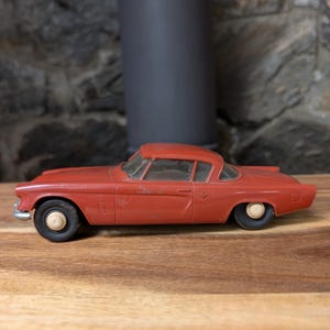May include: A vintage, red toy car with black tires and white hubcaps. The classic car model is a coupe with a detailed body and windows. The toy car is made of metal and sits on a wooden surface.