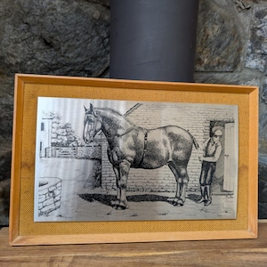May include: Framed black and white etching of a horse and a person. The horse stands before a brick building, with a person beside the door. The frame is wooden, with a burlap-like texture. The image is in a rectangular shape.