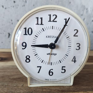 May include: White analogue alarm clock with black hands and numbers. The clock face reads "Kresfal Westclox" and "Made in U.S.A."