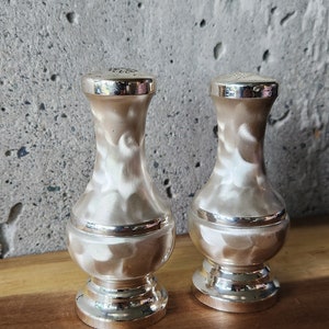 May include: A pair of silver salt and pepper shakers with a white swirl design. The shakers have a round base and a narrow neck.