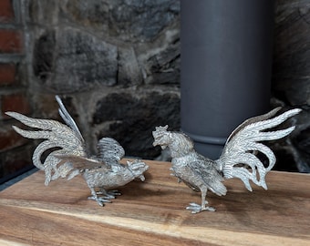 May include: Two silver rooster figurines with detailed feather designs stand on a wooden surface. The roosters face each other, showcasing their intricate craftsmanship. The background features a stone wall and a dark metal pipe.