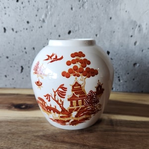 May include: A white ceramic vase with a brown and orange floral design. The vase features a traditional Chinese-inspired pattern with a pagoda and trees.