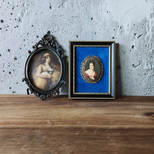 May include: Two antique oval-shaped picture frames with ornate gold metal frames. The frames are hanging on a gray concrete wall. The frames contain portraits of women in vintage clothing.