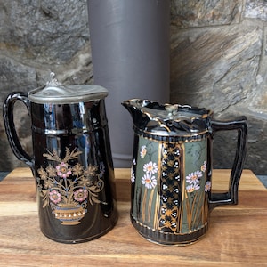 May include: Two antique black ceramic pitchers with ornate floral designs. One pitcher has a silver lid and a handle. The other pitcher has a green and gold floral pattern with a handle. Both pitchers are on a wooden surface.