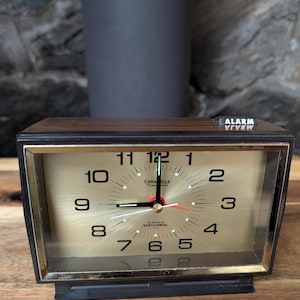 May include: A vintage Caraelle alarm clock with a gold face, black numbers, and a red second hand. The clock has a dark brown wood-grain finish and a black base. The word "ALARM" is visible on the top.