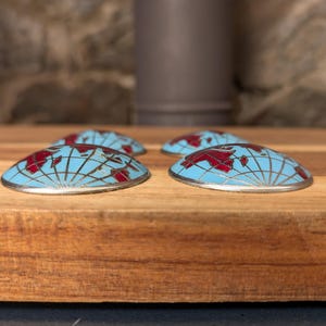 May include: Four decorative, half-dome shaped objects with a world map design. The objects have a light blue background with a red map and silver trim. They are arranged on a wooden surface.