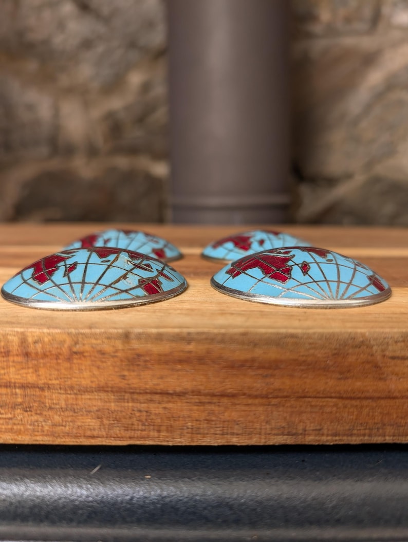 May include: Four decorative, half-dome shaped objects with a world map design. The objects have a light blue background with a red map and silver trim. They are arranged on a wooden surface.