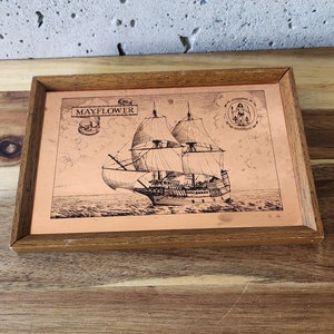 May include: A framed copper plaque with a black and white illustration of the Mayflower ship sailing on the ocean. The plaque has a wooden frame and the text "MAYFLOWER" is visible at the top.